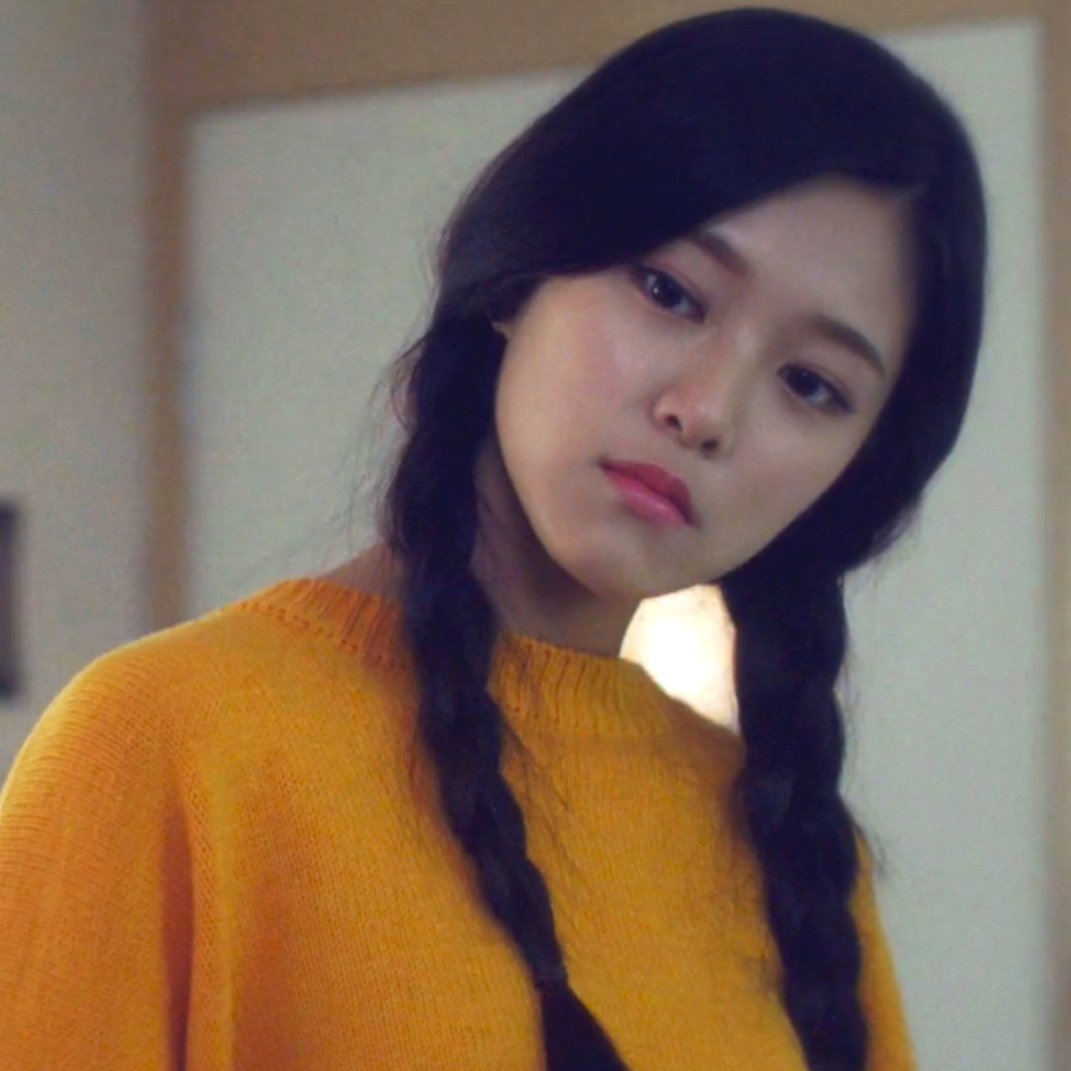 Hyunjin from the 'Around You' music video. She has long black hair tied into pigtails and is wearing a yellow sweater. She is tilting her head in curiosity.